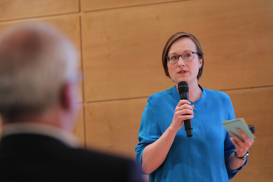 Eine Frau mit Brille und blauer Bluse hält ein Mikrofon in der Hand und spricht.