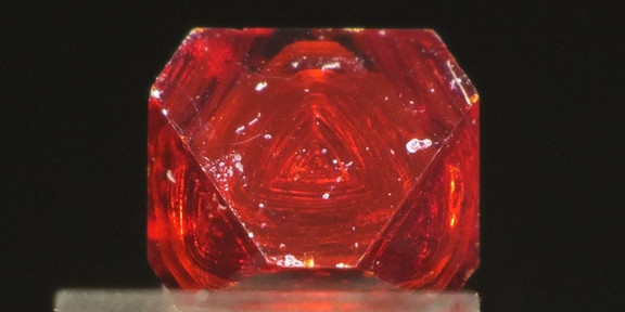 Foto: Ein roter, transparenter Kristall steht auf einer hellen Unterlage vor dunklem Hintergrund.