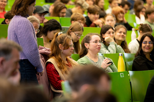 Auf dem Foto sieht man viele Personen im Hörsaal sitzen. Im Fokus ist eine Frau, die ein Mikrofon in der Hand hält und spricht.