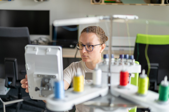 Eine Frau mit Brille sitzt an einer Stickmaschine und schaut konzentriert auf den Bildschirm der Maschine. Im Vordergrund sind mehrere farbige Garnspulen zu sehen.