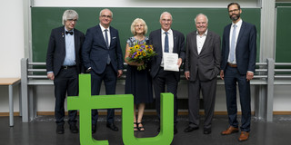 Six people in suits stand behind the green TU logo in front of a blackboard.