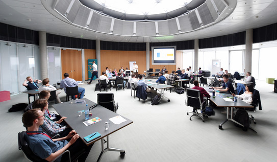 Konferenzsaal mit zehn verteilten Tischen, an denen kleinere Studierendengruppen sitzen und auf eine Beamerwand schauen.