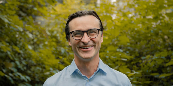 Porträtfoto von Benedikt Sabaß: Prof. Sabaß trägt ein hellblaues Hemd und lacht in die Kamera. Im Hintergrund sind Büsche.