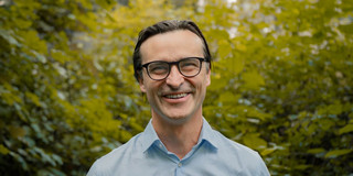 Porträtfoto von Benedikt Sabaß: Prof. Sabaß trägt ein hellblaues Hemd und lacht in die Kamera. Im Hintergrund sind Büsche.
