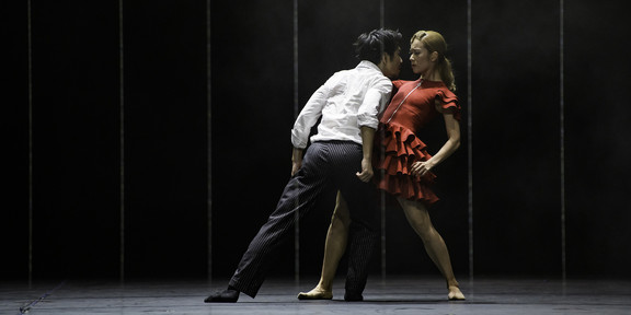 Foto: Ein Tänzer in Hemd und dunkler Hose und eine Tänzerin im roten Kleid stehen gemeinsam auf der Bühne in der Ballett-Produktion "Carmen".