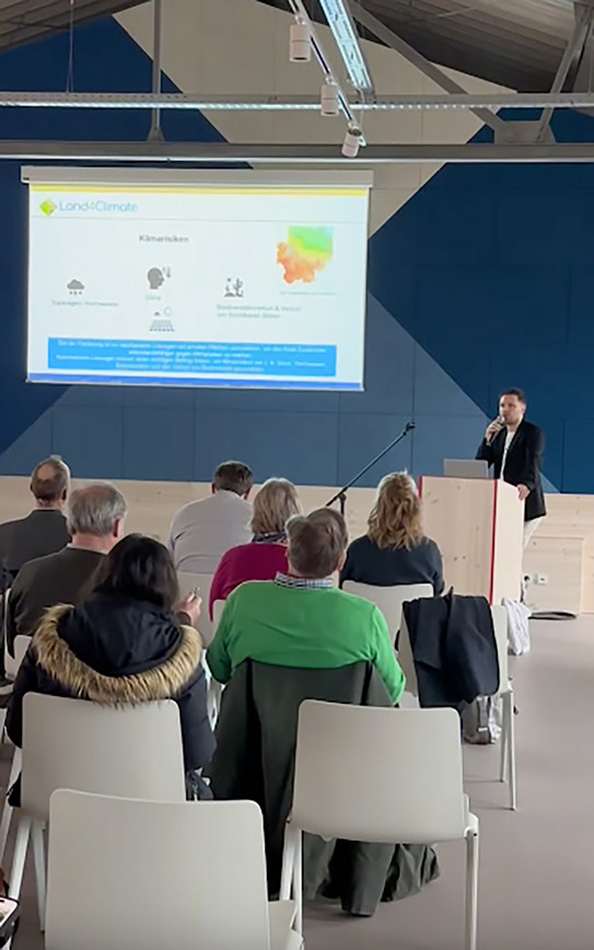 Foto: Eine Person hält einen Vortrag vor einem Publikum, während eine Präsentation zum Thema Klima auf einer Leinwand gezeigt wird.