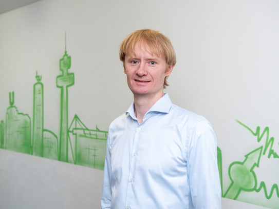 Portrait photo of Dr. Alex Greilich in front of a white wall with a green Dortmund graffiti skyline