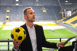 Christoph Edeler, Leiter des Hochschulsports, hält einen signierten Fußball im schwarz-gelben BVB-Design in seiner rechten Hand und moderiert. Im Hintergrund das Innere des Fußballstadions..