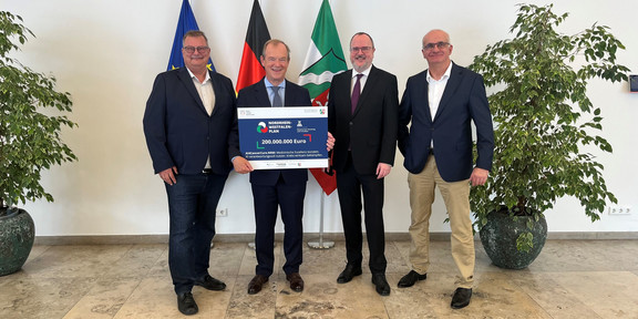 Foto: Vertreter*innen des Projekts präsentieren gemeinsam den Förderbescheid vor den Flaggen von Europa, Deutschland und Nordrhein-Westfalen.