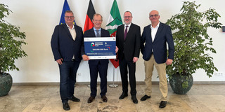 Foto: Vertreter*innen des Projekts präsentieren gemeinsam den Förderbescheid vor den Flaggen von Europa, Deutschland und Nordrhein-Westfalen.