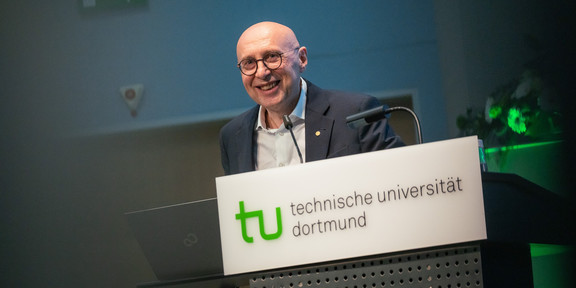Foto: Eine Person steht am Pult der Technischen Universität Dortmund mit Laptop und Mikrofon, davor ist ein Blumenarrangement zu sehen.