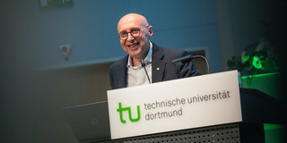 Foto: Eine Person steht am Pult der Technischen Universität Dortmund mit Laptop und Mikrofon, davor ist ein Blumenarrangement zu sehen.