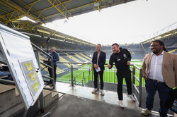Kevin Großkreutz, der auf der Tribüne des BVB-Stadions mit Darts-Pfeilen auf ein Whiteboard zielt. Neben ihm stehen drei weitere Männer.
