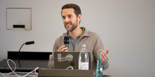 Auf dem Foto sieht man einen Gastredner in ein Mikrofron sprechen, der an einem Pult steht.