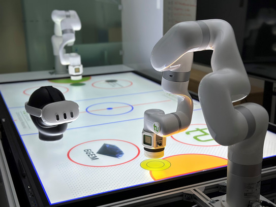 Zwei weiße Roboterarme interagieren mit einem beleuchteten, interaktiven Tisch. Auf der Oberfläche sind farbige Kreise und Symbole zu sehen. Daneben liegt ein VR-Headset; im Hintergrund ist ein weiterer Roboterarm sichtbar.
