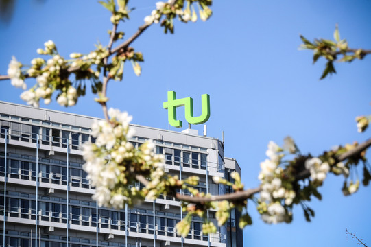 Das TU-Logo des Mathetowers vor einem wolkenlosen blauen Himmel, im Vordergrund ist leicht unscharf ein blühender Zweig eines Kirschbaums zu sehen.