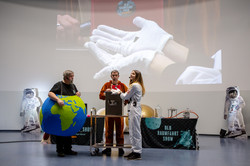 Drei Personen stehen in einem Hörsaal. Einer trägt ein Kostüm der Erde, ein weiterer ein Känguru-Kostüm. Eine Frau in einem Astronauten-Kostüm hält etwas in der Hand.