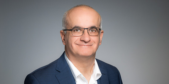 Porträtfoto von Prof. Manfred Bayer: Prof. Bayer trägt einen Anzug und eine Brille und steht vor einem grauen Hintergrund. Er lächelt in die Kamera.