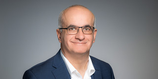 Porträtfoto von Prof. Manfred Bayer: Prof. Bayer trägt einen Anzug und eine Brille und steht vor einem grauen Hintergrund. Er lächelt in die Kamera.