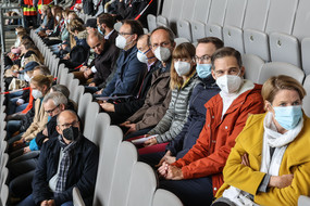 Es sitzen Menschen verteilt auf mehrere Reihen auf einer Tribüne in einem Fußballstadion und tragen eine Mund-Nasen-Bedeckung.