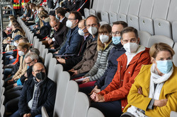 People are sitting in several rows in the stands of a soccer stadium and are wearing mouth-to-nose coverings.