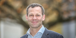 Portrait of Juniorprof. Wolfram Helml: An approximately 40-year-old man with short, light brown hair looks into the camera with a smile. He is wearing a gray jacket and a light blue and white checkered shirt.... 