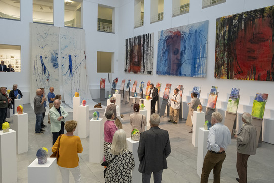 Mehrere Menschen bei einer Vernissage. In der Mitte des Raumes stehen Kunstwerke auf Säulen, an den Wänden hängen große Gemälde.