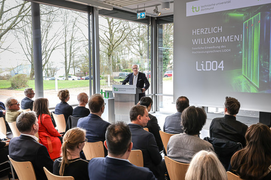 Foto: Eine Person spricht vor Publikum an einem Rednerpult, während eine Präsentationsfolie gezeigt wird.