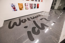 Auf dem Boden eines Ausstellungsraums steht "Warum ich?", an der Wand hängen Fotos