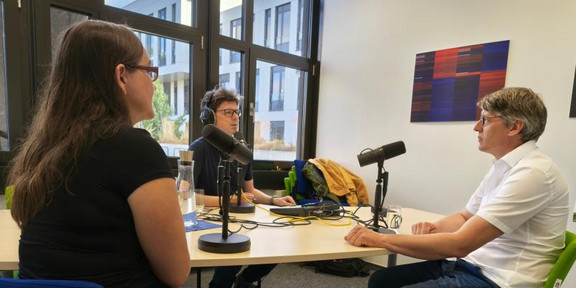 Eine Frau und ein Mann sitzen mit einem weiteren Mann an einem Tisch und nehmen einen Podcast auf.
