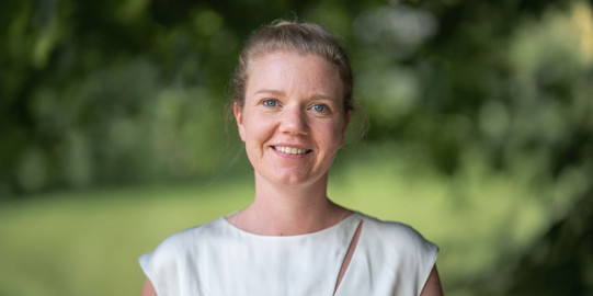 Landscape portrait photo of Prof. Kirsten Schorning: Prof. Schorning is wearing a white top and smiling at the camera. She is standing in front of a blurred green background.