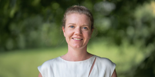 Porträtfoto von Prof. Kirsten Schorning im Querformat: Prof. Schorning trägt ein weißes Oberteil und lächelt in die Kamera. Sie steht vor einem verschwommenen grünen Hintergrund.