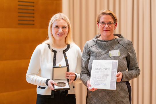 Zwei Frauen stehen nebeneinander. Die linke Frau hält eine Schachtel, in der sich eine Medaille befindet, die rechte Frau hält eine Urkunde in ihren Händen.