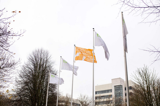 Fünf Fahnen an Fahnenmasten. Vier Fahnen sind weiß mit TU Dortmund Logo. Die mittlere Fahne ist orange.