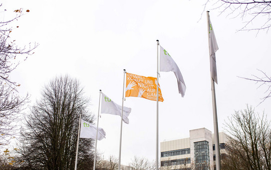 Fünf Fahnen an Fahnenmasten. Vier Fahnen sind weiß mit TU Dortmund Logo. Die mittlere Fahne ist orange. 