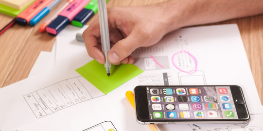 Jemand notiert etwas auf PostIT, daneben liegen Zettel und Smartphone