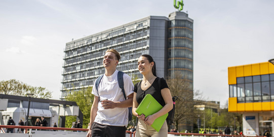 Zwei Studierende laufen nebeneinander über die Mensabrücke, im Hintergrund der Mathetower im TU-Logo