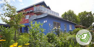 Blick auf ein Wohnhaus, das zum Teil einen roten Anstrich, zum anderen Teil einen blauen Anstrich hat. Im Vordergrund sind viele Pflanzen zu sehen.