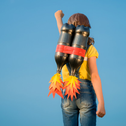 You can see a young person with a yellow T-shirt, jeans and leather cap from behind against a blue sky. She has a mockup of a rocket backpack on her back and stretches her left fist in the air.