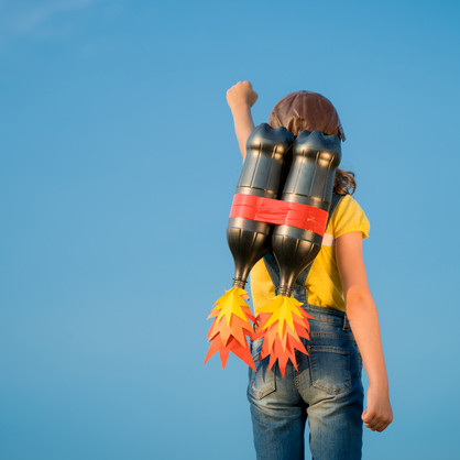 Man sieht eine junge Person mit gelbem T-Shirt, Jeans und Lederkappe von hinten vor einem blauen Himmel. Sie hat eine Attrappe eines Raketenrucksacks auf dem Rücken und streckt die linke Faust in die Höhe.
