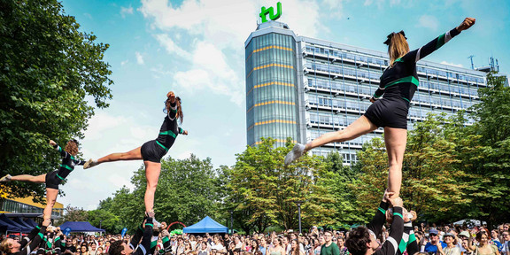 Zu sehen ist der Auftritt einer Cheerleader-Gruppe. Drei Chearleaderinnen werden von je drei Personen in die Luft gehoben. Im Hintergrund stehen viele Menschen.