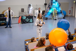 Eine Frau steht in einen vollbesetzten Hörsaal. Kinder die im Publikum sitzen tragen Wasserbälle, die verschiedene Planeten symbolisieren.