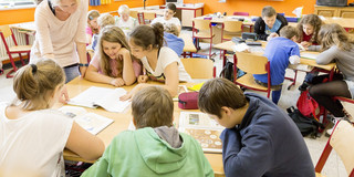 Mehrere Kinder sitzen an Tischgruppen in einem Klassenraum und lesen in Schulbüchern. Eine Lehrerin steht neben zwei Kindern und schaut mit ihnen in ein Buch.