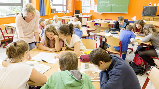 Mehrere Kinder sitzen an Tischgruppen in einem Klassenraum und lesen in Schulbüchern. Eine Lehrerin steht neben zwei Kindern und schaut mit ihnen in ein Buch.