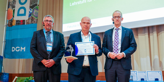 Drei Männer vor einer Präsentation. Der Preisträger Prof. Walter steht in der Mitte und hält Pokal und Urkunde in den Händen.