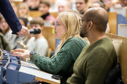 A man and a woman are sitting in the lecture hall. The woman asks a question into the microphone.
