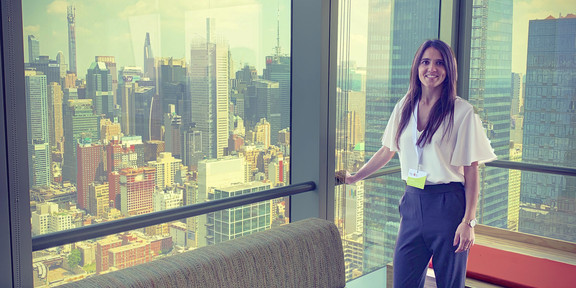 Eine Frau steht an einem Panoramafenster, im Hintergrund sind Gebäude in New York City zu sehen