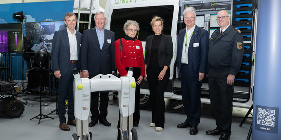 Mehrere Menschen vor einem Exponat auf dem Logistik Campus Hinter einem Exponat auf dem Logistik Campus stehen Mona Neubaur, Dirk Aschenbrenner von der Feuerwehr Dortmund und dem Deutschen Rettungsrobotikzentrum, Prof. Wietfeld, Monika Gatzke vom CC5G.NRW, Prof. ten Hompel und Prof. Henke, beide vom Fraunhofer IML und der TU Dortmund).