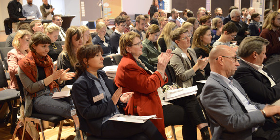 Several people sit in the audience during a meeting and clap.