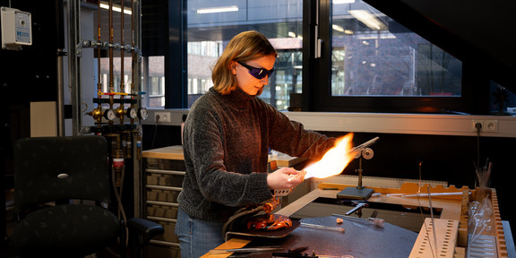 Foto: Eine Person arbeitet in einem Labor mit einer Flamme und verschiedenen Glasinstrumenten.